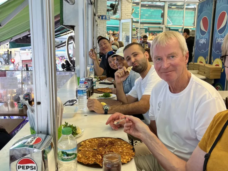 Guests along the counter of a lahmacun shop, eating and turning to the camera