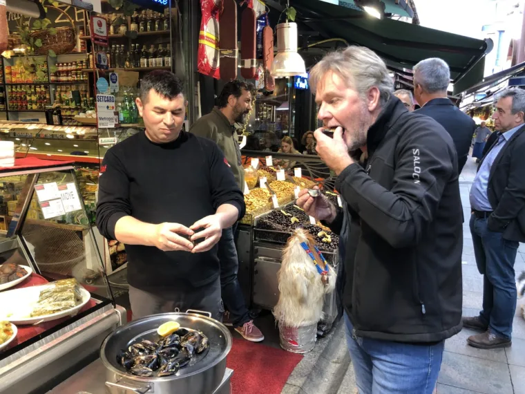 A guest tasting at a delicatessen counter while the shopkeeper serves