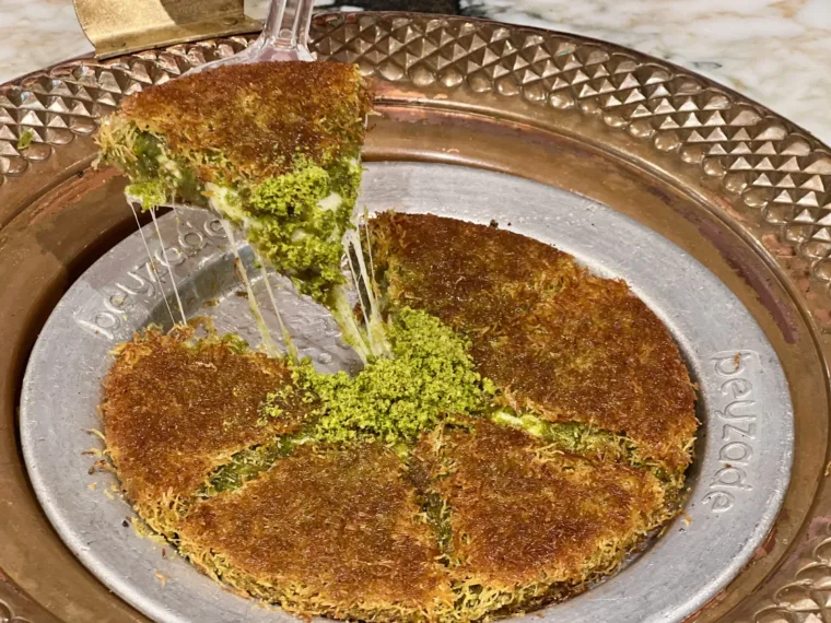 Kunefe pulled apart on a copper plate, the cheese stretching under crushed pistachio